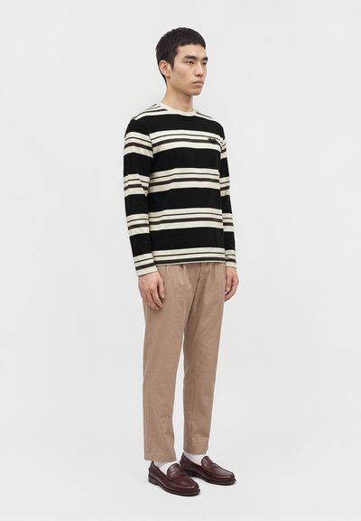 Homme debout, face à droite, portant une chemise rayée noir et beige à manches longues, un pantalon beige, des chaussettes blanches et des mocassins marron foncé sur un fond uni.