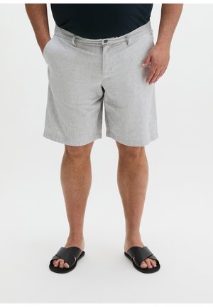 Man draagt lichtgrijze knielange shorts en zwarte slip-on sandalen, staand met handen in de zakken op een witte achtergrond.