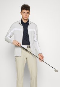 Chaqueta gris claro con cremallera y rayas blancas, polo azul marino, pantalones beige y un palo de golf. Presenta una tela suave y un corte moderno.