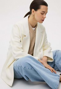 Overmaatse witte jas over een beige trui en lichtblauwe brede jeans. Gladde stof met gestructureerde revers en grote knopen.