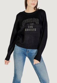 Schwarzer Pullover mit "GUESS LOS ANGELES"-Schriftzug und Strassverzierung, lockerer Schnitt, weite Ärmel, kombiniert mit hellblauen Jeans.