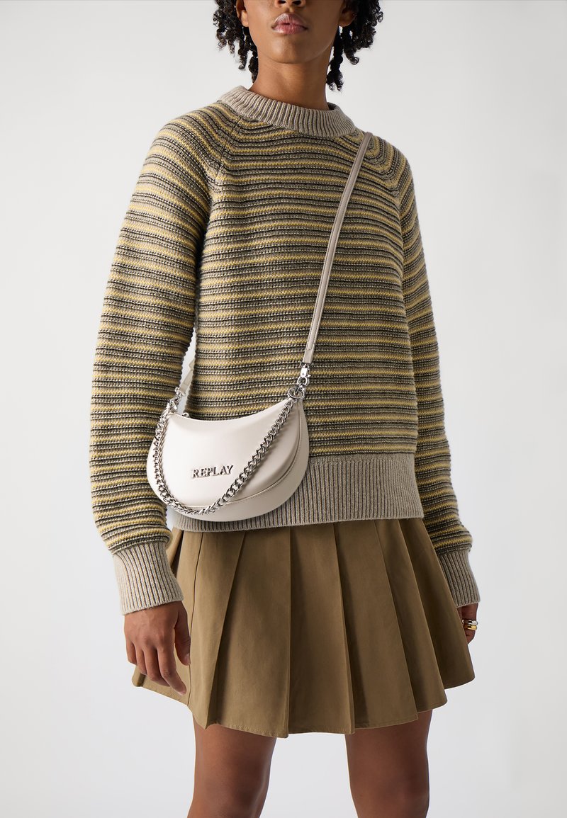 Sac en cuir gris clair porté en travers avec une chaîne en argent, présentant le mot "REPLAY" en relief. Associé à un pull en tricot rayé et une jupe plissée.