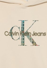 Felpa beige con il testo "Calvin Klein Jeans" in verde, e un grande motivo "CK" integrato in un design di edificio. Texture morbida.