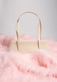 Sac à main rectangulaire beige avec double anse reposant sur un tissu rose doux et duveteux, sur un fond gris clair.