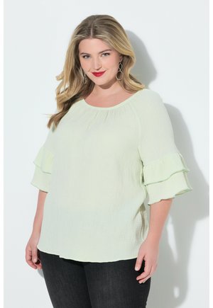 Lachende vrouw met blond haar, hangende oorbellen, een lichtgroene blouse met ruches aan de mouwen en een zwarte broek, voor een witte achtergrond.