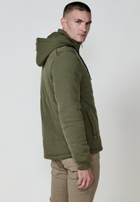 Žalia siuvinėta zip-up gobtuvinė striukė su raišteliu, tekstūruota medžiaga ir šoniniais kišenėmis, dėvima su smėlio spalvos kelnėmis, rodomas iš šono.