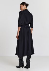 Frau mit einem Dutt trägt ein schwarzes, langärmliges Midikleid mit geraffter Taille, kombiniert mit schwarzen spitzen, mittelhohen Stiefeln und steht mit dem Rücken zur Kamera.
