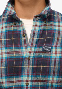 Sininen ja ruudullinen flanellipaita, jossa on punaisia, vihreitä ja keltaisia ruutuja, napit sulkemiseen ja rinnataskussa logopainatus.