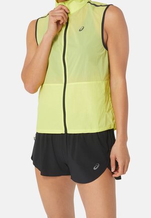 Osoba ubrana w żółtą kamizelkę na zamek bez rękawów oraz czarne krótkie spodenki sportowe z logo, stojąca na tle jasnego, gładkiego tła.