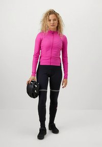 Dopasowana różowa kurtka kolarska z przednim zamkiem; zestawiona z czarnymi ocieplanymi legginsami kolarskimi z odblaskowymi akcentami. W ręku trzymany jest czarny kask.