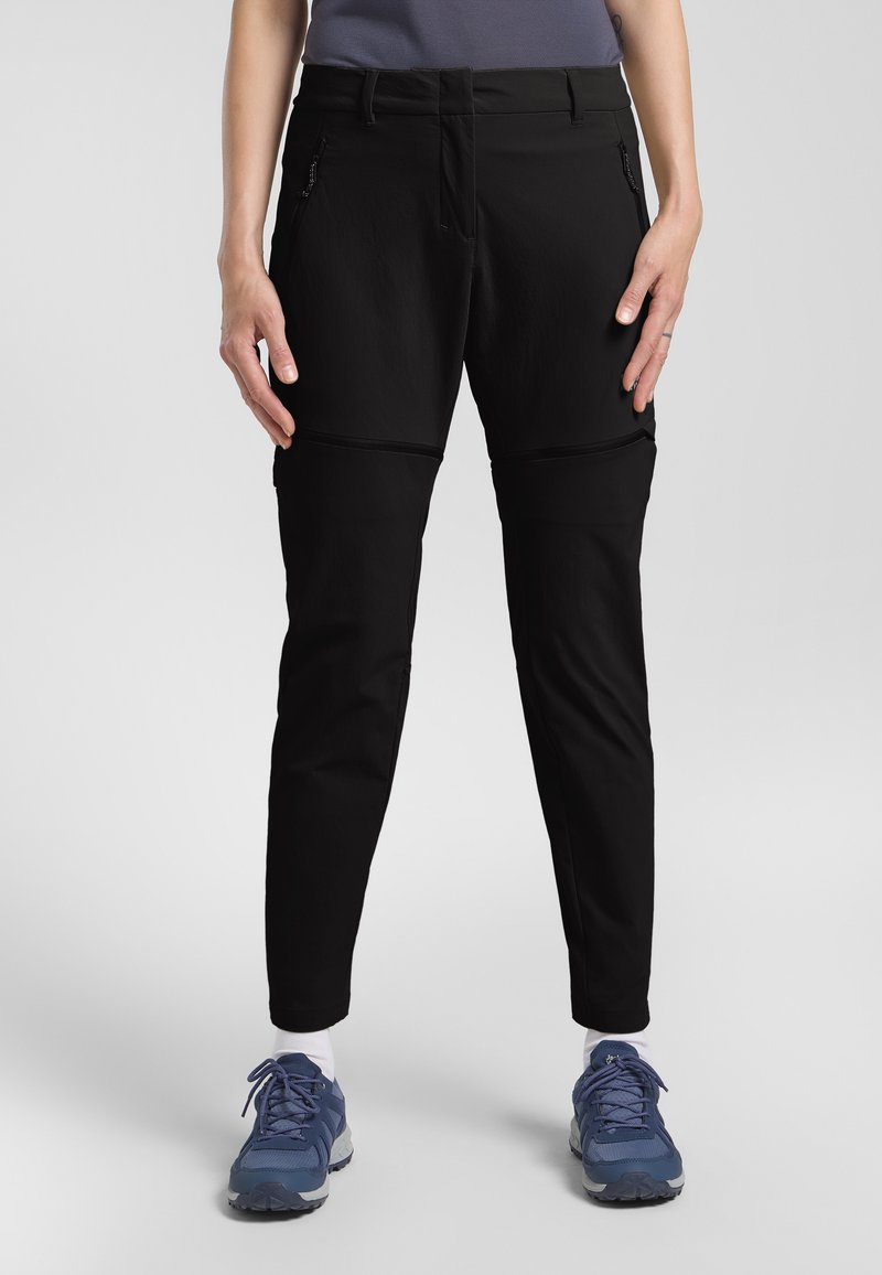 Pantalons noirs fuselés avec une texture lisse, dotés de poches zippées et de coutures renforcées. Portés avec des chaussures de sport bleues pour un contraste.