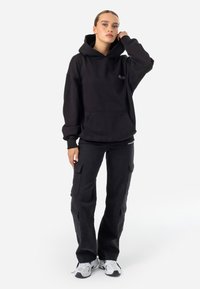 Felpa nera in cotone con grande tasca frontale e logo. Abbinata a pantaloni cargo neri con tasche laterali. Sneakers bianche con dettagli neri.