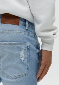 Světlé modré džíny z denimu s potrhanými detaily a hnědou koženou nášivkou v pase. Ruka opřená o bok, ve spojení s bílou mikinou.