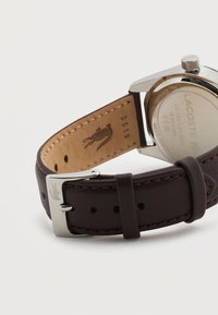 Καφέ δερμάτινο λουράκι ρολογιού με ασημένιο αγκράφα, χαραγμένο με λογότυπο κροκόδειλου, προσαρτημένο σε ασημένια σιλβεράτη πίσω θήκη ρολογιού από ανοξείδωτο χάλυβα.