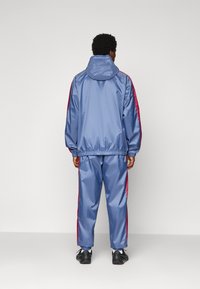 Mann, der einen blauen Kapuzen-Trainingsanzug mit roten Seitenstreifen und schwarzen Sneakers trägt, steht mit dem Rücken zu einem schlichten Hintergrund.