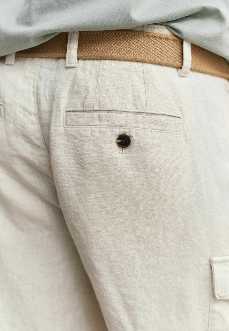 Pantalones de lino beige con un bolsillo trasero abotonado y cinturón trenzado color coñac, combinados con una camisa gris claro metida por dentro.
