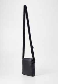 Crna crossbody torba napravljena od glatke kože, s pravokutnim oblikom, podesivim tekstilnim remenom i zatvaranjem na patentni zatvarač na vrhu.