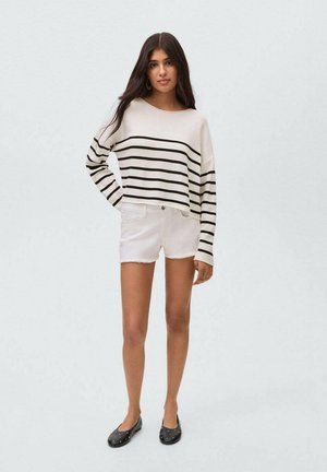 Gestreifter, weiß und schwarz, kürzer geschnittener Pullover, weiße, ausgefranste Denim-Shorts, schwarze Ballerinas mit silbernen Nieten. Das Model steht mit einer Hand an der Hüfte.