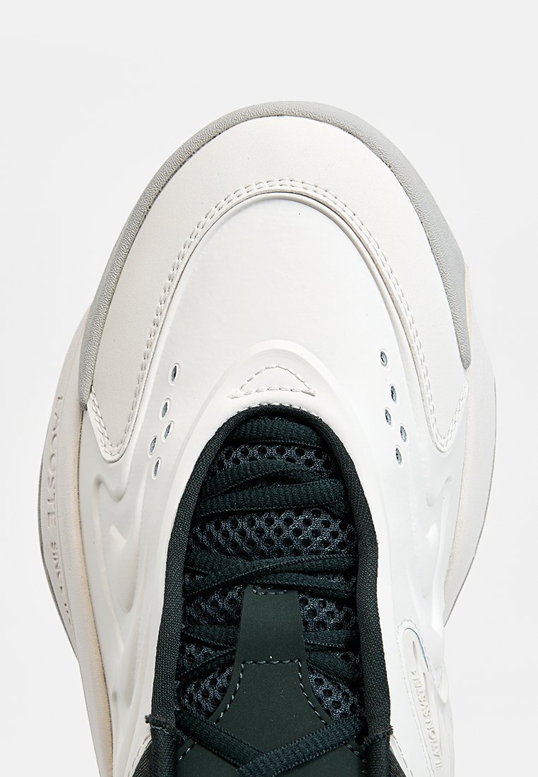 White leather sneaker featuring a padded gray collar, black mesh tongue, intricate lace details, and perforated accents on the sides.