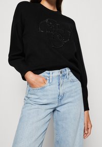 Suéter negro de punto con un diseño de logo texturizado en rhinestones negros, combinado con jeans de tiro alto de color azul claro. Tela suave, corte holgado.
