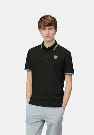 Giovane uomo che indossa una polo nera con bordi bianchi e un piccolo logo sul petto, in piedi con le mani nelle tasche di pantaloni grigio chiaro su sfondo bianco.