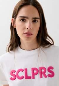 Bílý bavlněný tričko s velkým růžovým vyšitým textem "SCLPRS." Modelka nosí barevný korálkový náhrdelník a má rovné hnědé vlasy.