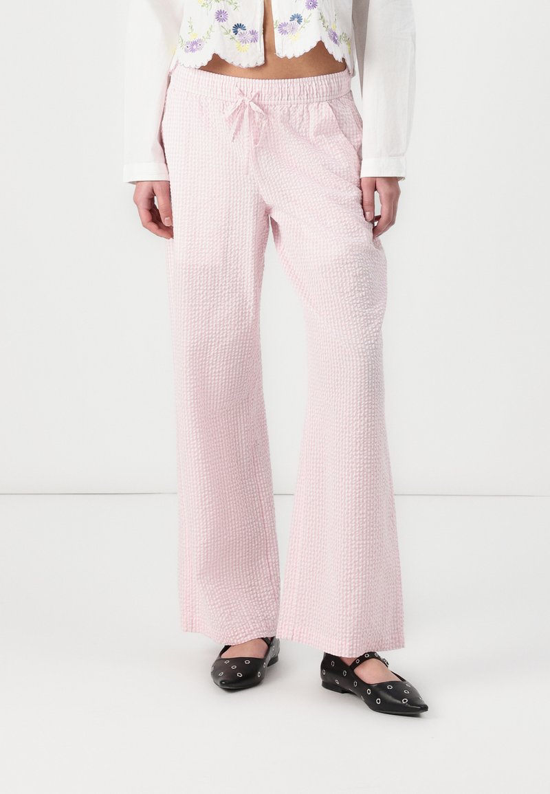 Pantalons larges rose clair, texturés, avec une taille élastique et un nœud à l'avant. Portés avec des chaussures plates noires à accents circulaires.