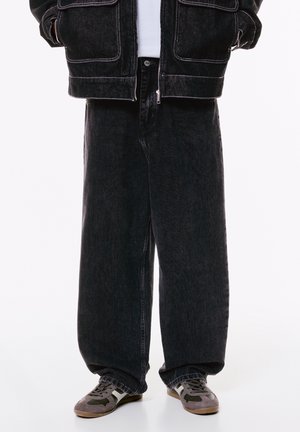 Personne portant un jean noir ample, une veste en denim noire avec des surpiqûres blanches et des baskets grises avec des bandes blanches.