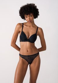 Melna mežģīņu bralette un thong komplekts ar sarežģītiem rakstiem, viļņotiem galiem un plānām siksnām, uzsverot delikātu, teksturētu dizainu.