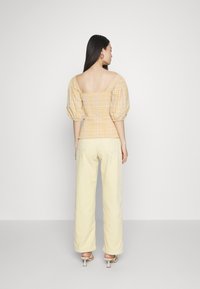 Top amarelo à quadrados com mangas bufantes, decote quadrado e detalhes elásticos, combinado com calças largas amarelas pálidas. Sapatos de salto fino.
