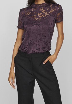 Kvinde iført en kortærmet lilla bluse i blonder med blomsterprint, som er stoppet ned i højtaljede sorte bukser, med hånden i lommen.