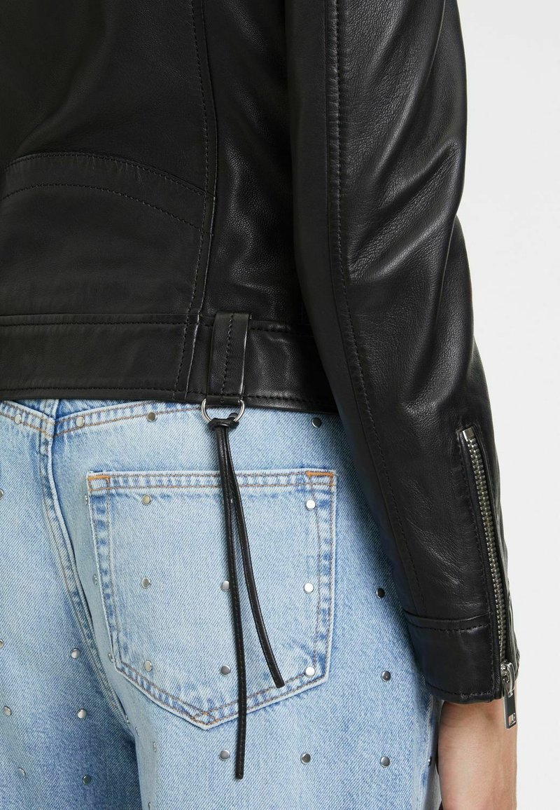 Veste en cuir noire à texture lisse, avec des manches zippées et un cordon décoratif à l'ourlet, associée à un denim bleu clair clouté.