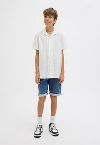 Camicia bianca a maniche corte con texture e bottoni, abbinata a un paio di pantaloni shorts in denim arrotolati e sneakers bianco/nero, su uno sfondo chiaro.