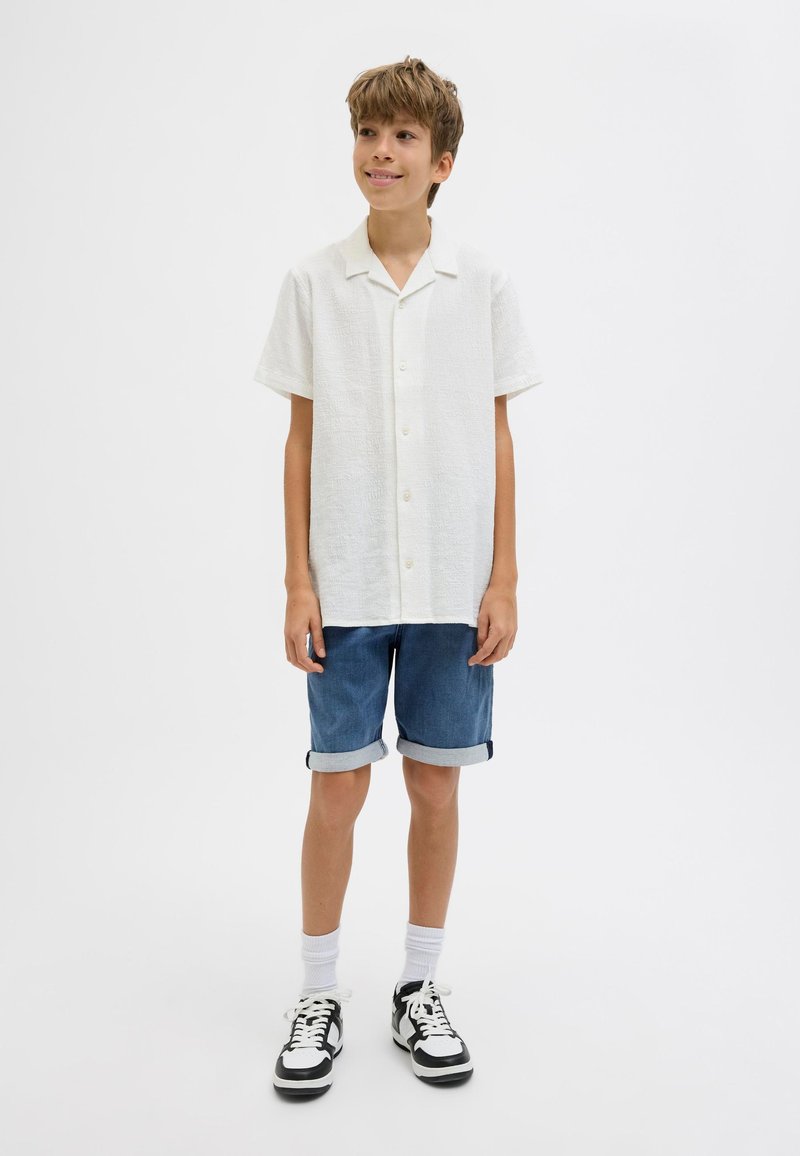 Camicia bianca a maniche corte con texture e bottoni, abbinata a un paio di pantaloni shorts in denim arrotolati e sneakers bianco/nero, su uno sfondo chiaro.