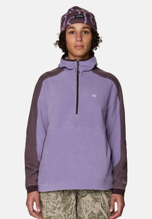 Person trägt eine violette und braune Fleecejacke mit Viertelreißverschluss, eine violette gemusterte Mütze und grüne Tarnhose und steht vor einem schlichten Hintergrund.