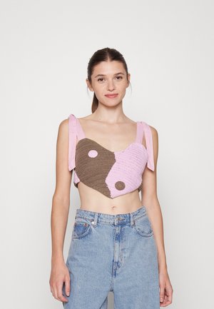Top cropped em croché nas cores rosa e castanho, apresentando uma forma de coração, largas alças rosas e dois detalhes circulares. Combinado com jeans de cintura alta.