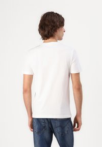 Valkoinen puuvillapaita, jossa on pyöreä kaula-aukko ja lyhyet hihat, käytössä henkilö, joka on selkä kameraan. Denim-farkut näkyvät vyötäröllä.