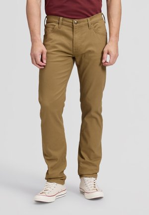 Homme portant un pantalon couleur moutarde et des baskets blanches, debout devant un fond uni, les mains détendues le long du corps.