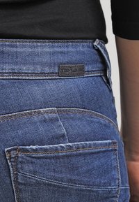 Jean fabric in dark blue with a smooth texture, featuring a leather logo patch, back pockets, and contrast stitching details.