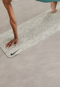 Colchoneta de yoga de Nike con una superfície texturizada de color gris claro con manchas negras, bordes redondeados y un logo negro en la parte inferior.