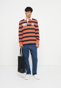 Camisa de rugby às riscas em laranja e azul marinho com colarinho branco, apresentando o número 85 e o emblema de um leão. Acompanhada de jeans azuis e uma mala tote preta.