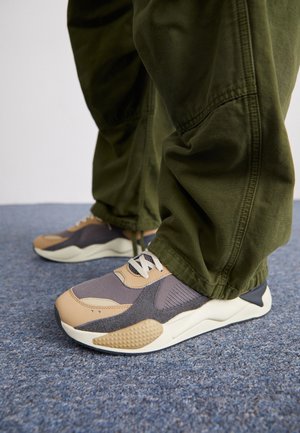 Persona que lleva zapatillas multicolor beige y grises con suelas blancas, de pie sobre una alfombra azul, vestida con pantalones sueltos de color verde oliva.