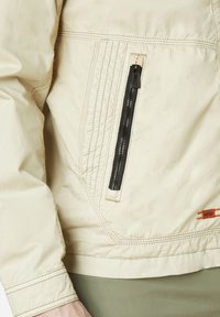 Beige lightweight jacket with a zippered pocket, visible stitching, and a textured finish. Features a logo in orange on the lower right.