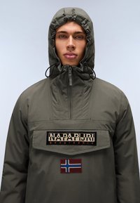 Aliejinės žalios spalvos striukė su gobtuvu ir užtrauktuku, turinti didelę priekinę kišenę, ženkliuką ir Norvegijos vėliavos detalę. Lygus, vandeniui atsparus audinys.