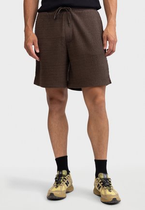 Homme portant un short marron à cordon texturé, des chaussettes noires et des chaussures de course jaunes sur un fond clair.