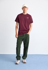 Burgundy cotton t-shirt with small white logo, paired with green corduroy pants and white sneakers with red accents, standing on a blue floor.