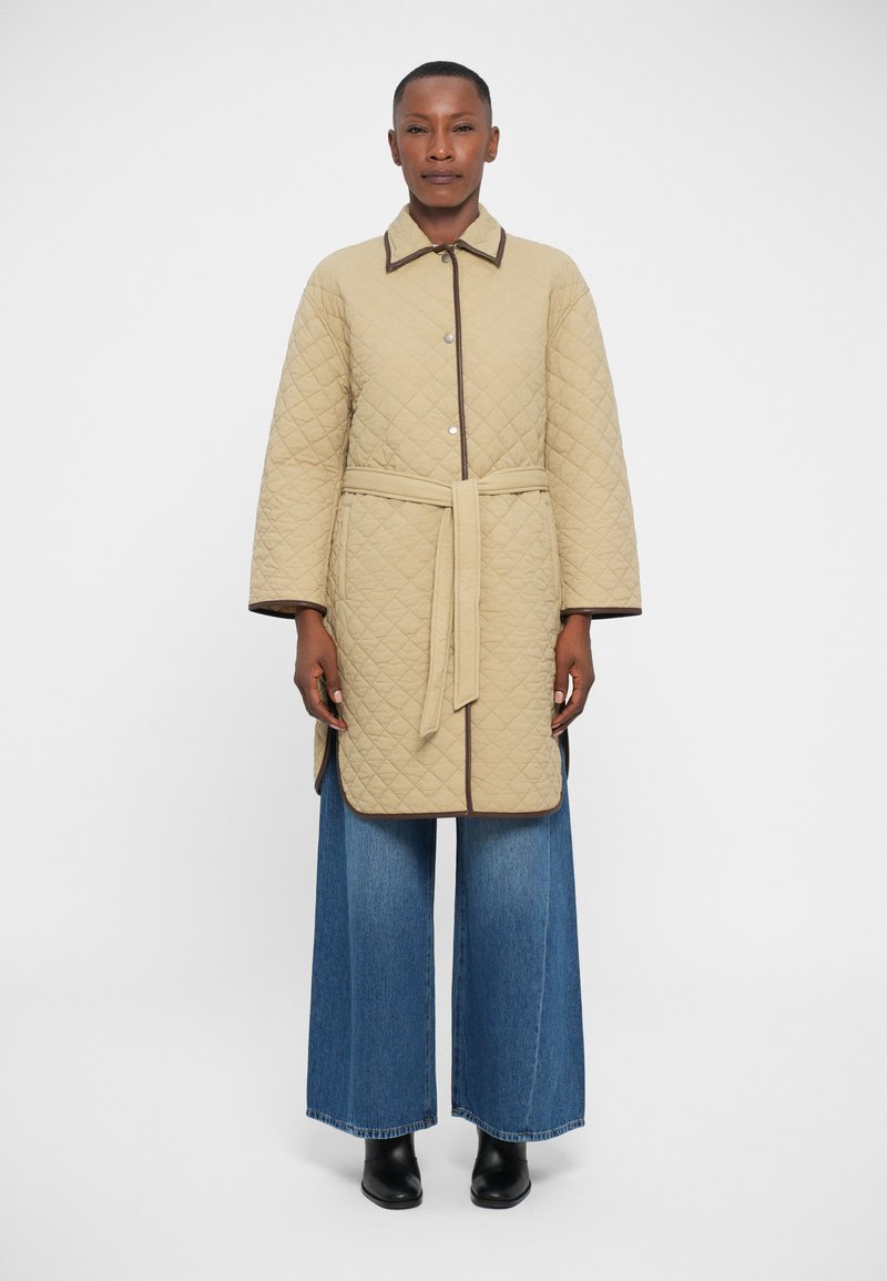 Quilted beige coat with a collar, button closure, and a tied belt. Contrast brown trim outlines the hem and sleeves. Wide-leg blue denim pants.