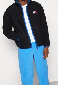 Persona che indossa una giacca nera zip con texture e rifiniture blu, insieme a pantaloni di pile azzurri, in piedi contro uno sfondo neutro.