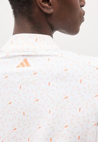 Άτομο που φοράει λευκό πουκάμισο με μικρά μοτίβα φύλλων ροδακινιού και λογότυπο Adidas ροδακινιού στο πάνω μέρος της πλάτης, γυρμένο προς τα δεξιά.