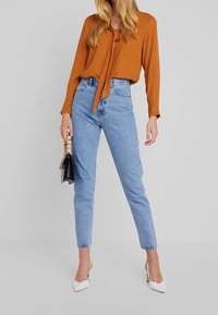 Brun bluse med sløjfedetalje, lyseblå højtaljede jeans og hvide spidse hæle. Sort håndtaske med træhåndtag.